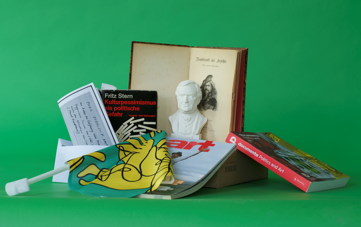 On a green surface lie 3 books, a letter, a magazine, a flag and a bust.