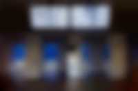 Ausstellungsansicht im Flur: 3 Pfeiler mit Spiegelfliesen verkleidet, im Hintergrund mit blauer Folie beklebte Glastüren, von der Decke hängen 2 Monitore mit einer Videoarbeit.