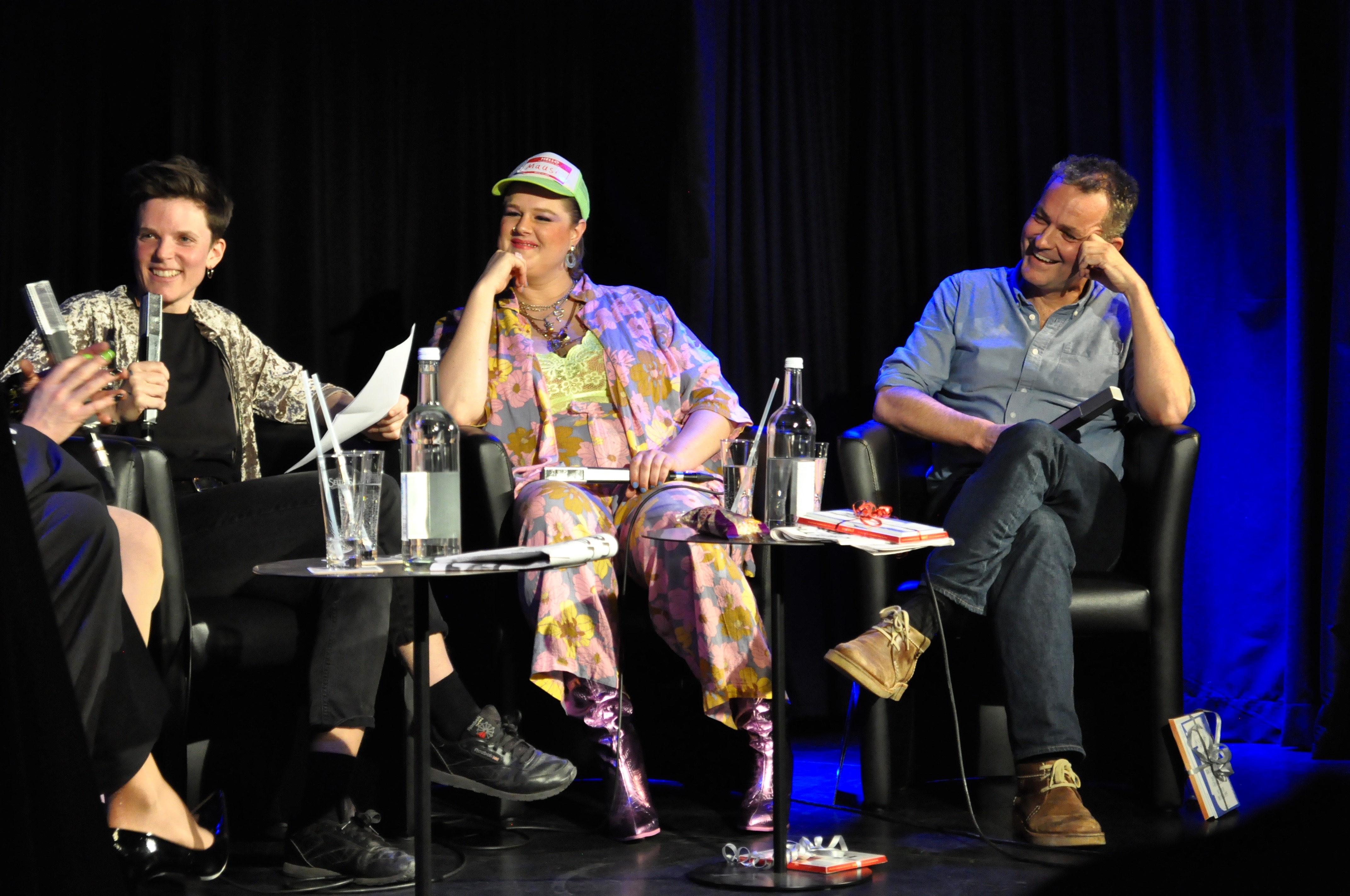 3 people with microphones sit on stage at the talk show and smile.