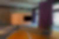 Ausstellungsansicht im Neubau: Von der Decke hängt ein Monitor, davor steht eine Bank auf einem orangenen Teppich, rechts hängt ein Vorhang in lila, im Hintergrund ist eine orangene Wand.